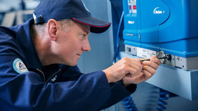 A technician in a blue uniform and cap working on machinery with a screwdriver.