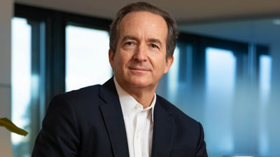 Um profissional de terno escuro e camisa branca, com uma expressão confiante e pensativa, sentado em um escritório com janelas grandes.