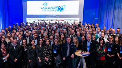 Un ampio gruppo di professionisti si è riunito a un evento aziendale, in piedi insieme sotto un cartello che recita “Il tuo successo, la nostra passione” in un locale illuminato di blu.