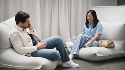 A imagem mostra dois colegas sentados em pufes confortáveis de cor clara em um ambiente informal e descontraído. O homem à esquerda, vestindo uma jaqueta leve e jeans, está segurando uma caneca e parece estar conversando. A mulher à direita, vestindo uma roupa azul-claro, está sorrindo e tem uma garrafa de água e uma bolsa bege ao seu lado. O fundo mostra uma cortina neutra, contribuindo para uma atmosfera aconchegante e casual.