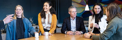 Six people engaged in a casual conversation around a wooden table in a modern office setting, with colourful artwork and hanging light bulbs in the background.