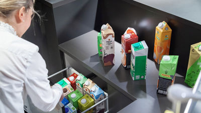 Profissional organiza diversas embalagens assépticas da Tetra Pak, com diferentes formatos e designs, em prateleira expositiva, evidenciando a variedade de soluções para cocriação de produtos, inovação em sistemas alimentares sustentáveis e envase seguro.