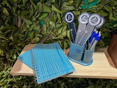 Brindes sustentáveis da Tetra Pak: cadernetas azuis e canetas em suporte com réguas personalizadas, todos feitos de material reciclado, expostos sobre prateleira com folhagens, parte da ação interativa com roleta para visitantes ganharem prêmios como o Caixito.