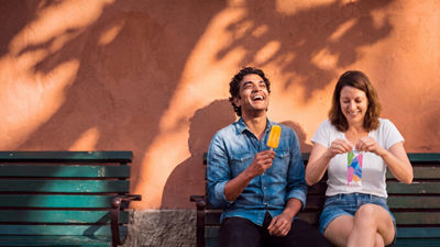 A man and a woman sit on a bench outdoors with ice lollies and colourful wrappers.