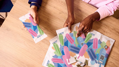 Hands examine colourful ice cream packaging samples on a wooden table.