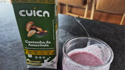 Caixa do produto Cuíca de Castanha da Amazônia ao lado de um copo de vidro com vitamina de cor roxa sobre uma bancada escura. A embalagem destaca que é um alimento à base de castanha-do-Brasil, com informações nutricionais e selo Tetra Pak