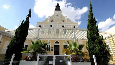 Fachada histórica da Santa Casa de Ponta Grossa, hospital referência em saúde, com torre de relógio central, estrutura amarela clássica, cobertura envidraçada e jardim com palmeiras e ciprestes, sob céu azul com nuvens.