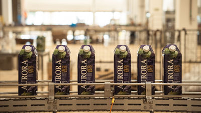 Seis caixas de suco de uva integral Aurora avançam por uma esteira industrial, ilustrando a parceria com a Tetra Pak para envase em embalagens cartonadas e maior eficiência operacional.