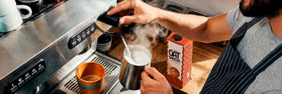 Barista steaming oat drink beside an espresso machine and oat barista carton