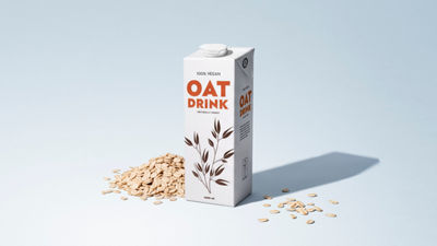 Carton of plant-based oat drink surrounded by oat flakes on a light blue background.