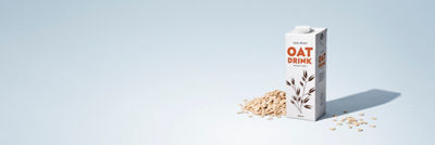 Carton of plant-based oat drink surrounded by oat flakes on a light blue background.