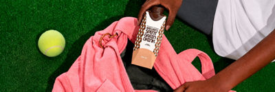 Person holding a carton of spent grain drink next to a pink sports bag and tennis ball on green grass.