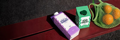 Cartons of plant-based blueberry yoghurt drink and soya drink next to a green shopping bag filled with oranges.