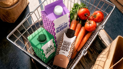 A shopping basket filled with fresh carrots, tomatoes and three colorful cartons of plant-based drinks, including soya, blueberry yoghurt-style and spent grain drinks.