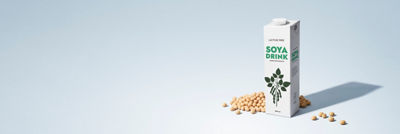 Carton of plant-based soya drink surrounded by soybeans on a light blue background.