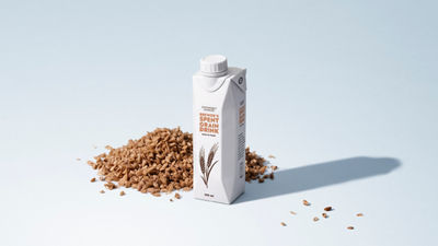 Carton of plant-based spent grain drink surrounded by spent grains on a light blue background.
