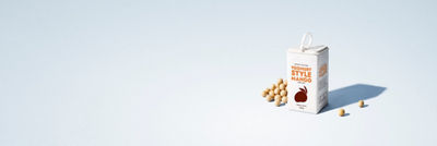 Carton of plant-based mango yoghurt-style drink made from soya, surrounded by soybeans on a light blue background.
