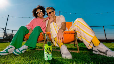 Man and woman sitting in beach chairs on the grass with an RTD alcoholic beverage in carton beside them