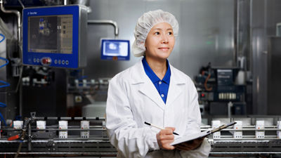 Employee wearing a white coat and hairnet holding a clipboard beside a carton filling line.
