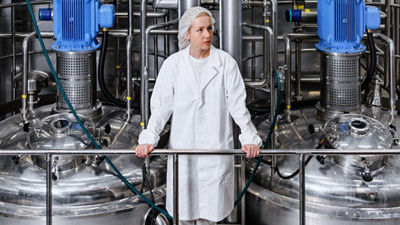 Employee wearing a white coat and hairnet standing between stainless steel tanks with blue motors.