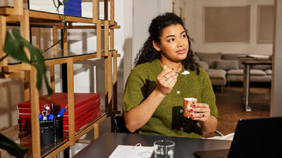 Person eating yoghurt at a desk while working on a laptop.