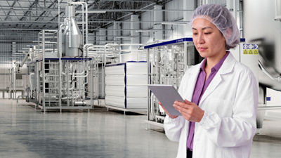Woman in a white coat and hairnet using a tablet beside processing equipment