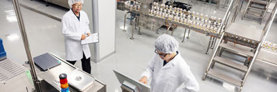 Two employees in white coats and hairnets beside a packaging line, one using a tablet