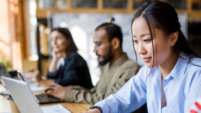 Tre persone lavorano in modo diligente sui laptop in uno spazio di lavoro moderno. L’attenzione è rivolta a una donna in primo piano con lunghi capelli scuri, che indossa una camicia blu, mentre due colleghi che lavorano alle rispettive mansioni sono sfocati sullo sfondo. L’ambiente sembra essere collaborativo e produttivo.