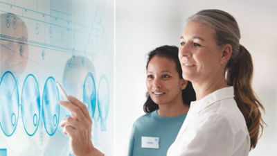 Two women collaborating at a digital whiteboard, one pointing with a pen at technical drawings while the other observes, both smiling and engaged in the discussion.