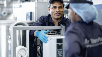 Funcionário da Tetra Pak de uniforme escuro e luvas azuis conversa com colega de trabalho que veste touca higiênica, em ambiente de produção industrial, em colaboração