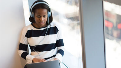 A person wearing headphones and a black-and-white striped sweater is sitting by a window and working on a laptop. The setting suggests a quiet and focused environment, possibly a study or work area.