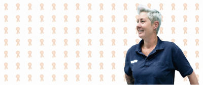 A person with short, light hair, dressed in a blue polo shirt, smiling against a background patterned with light beige ribbon icons, symbolising awards or recognition.
