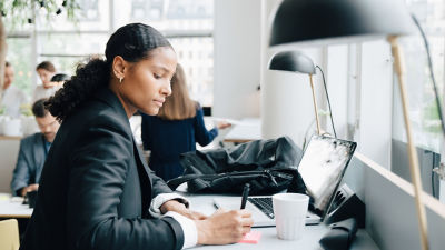 A professional woman with curly hair tied back is seated at a desk, writing notes. She is dressed in a black blazer, and beside her is a laptop and a cup, suggesting she is engaged in work or study. The setting appears to be a modern office or co-working space with other people in the background.