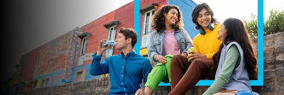 A group of young adults sitting together and chatting, with one person drinking from a carton, in a colourful outdoor urban area.