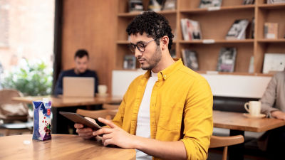 A man with curly hair and glasses is seated at a table in a café, reading from a tablet. He is wearing a yellow shirt over a white t-shirt. In front of him on the table is a carton of juice. In the background, there are shelves with books and another person working on a laptop.