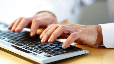 Close-up of a person's hands typing on a keyboard, focusing on the fingers and the keys. The individual appears to be wearing a white long-sleeve shirt, indicating a professional or office environment.