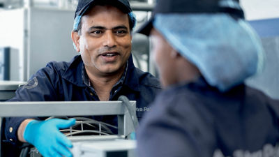 The image shows a man wearing a blue Tetra Pak uniform, cap, and hairnet, speaking and smiling at a colleague who is also wearing similar attire. They are in a clean, professional industrial environment, working with machinery. The man is wearing blue gloves, indicating a focus on hygiene and safety. The background includes industrial equipment, suggesting a manufacturing or production setting.