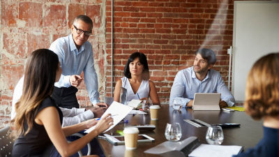 Un gruppo di professionisti in una sala riunioni con un grande tavolo in legno. Un uomo che indossa una camicia azzurra sta in piedi e parla, mentre altri, tra cui una donna che indossa una camicetta bianca e un uomo con un computer portatile, sono seduti e ascoltano con attenzione. La camera presenta una parete in mattoni a vista.