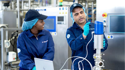 Two factory workers in blue uniforms and hairnets collaborating on a piece of equipment, with one explaining its function while the other observes and works on a laptop.