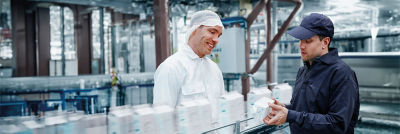 Three workers in a factory setting wearing hairnets and uniforms, discussing something on a mobile device next to a production line.