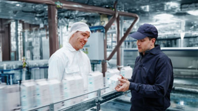 Two factory workers in a production line setting, one in a white hairnet and the other in a blue cap, inspecting a product and engaging in conversation amidst machinery and equipment.