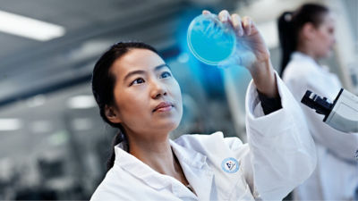 A woman in a lab coat holds an object up to inspect it closely.