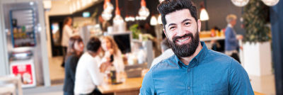 A bearded man with dark hair and a bright smile is wearing a blue button-up shirt in a cafe