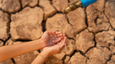 Cracked, dry earth with hands cupped below a water tap, with droplets coming out.