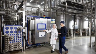 Two workers walking through an industrial processing facility with stainless steel equipment and pipelines