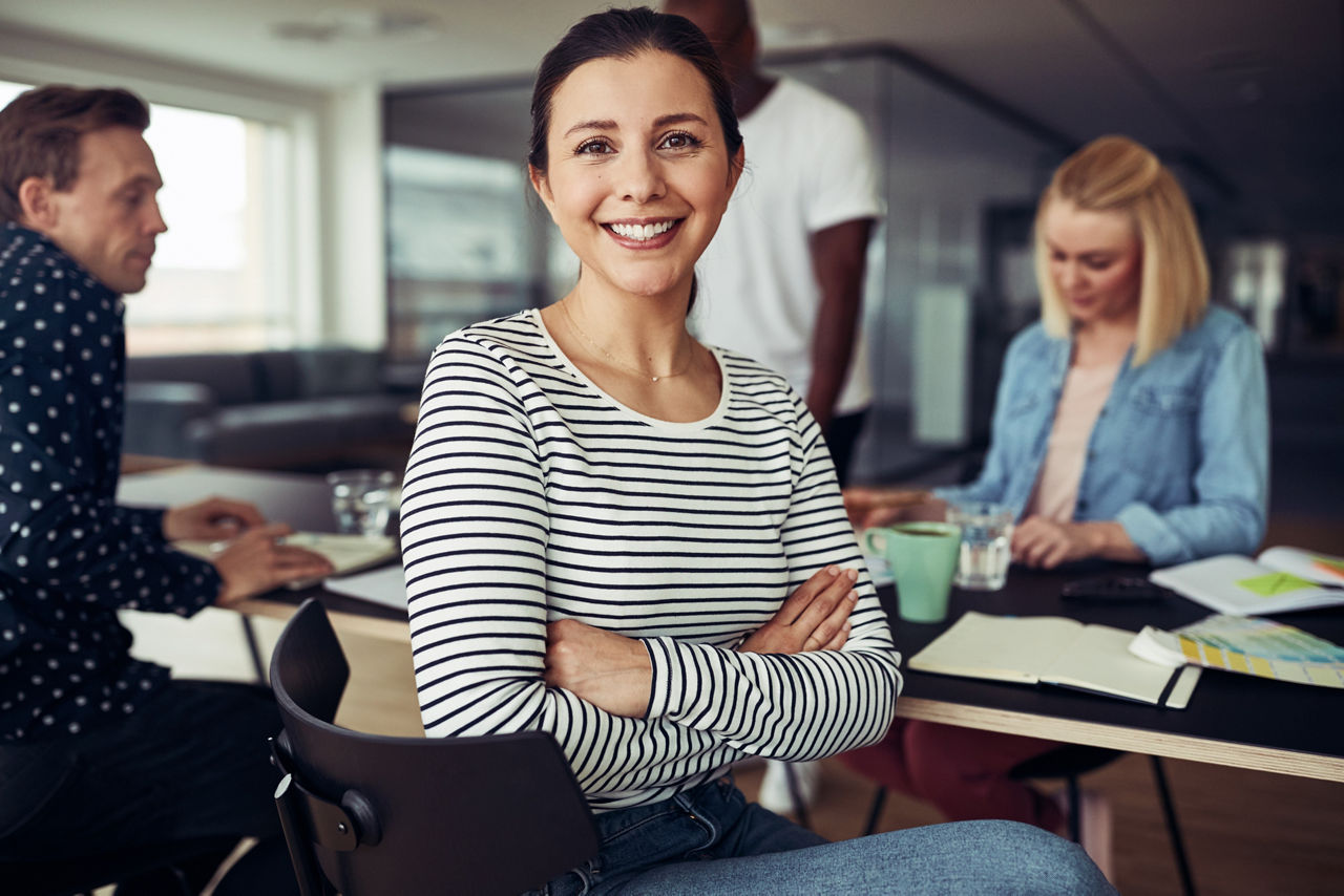 Young businesswoman smiling while sitting with her arms crossed at an office table with colleagues working in the background