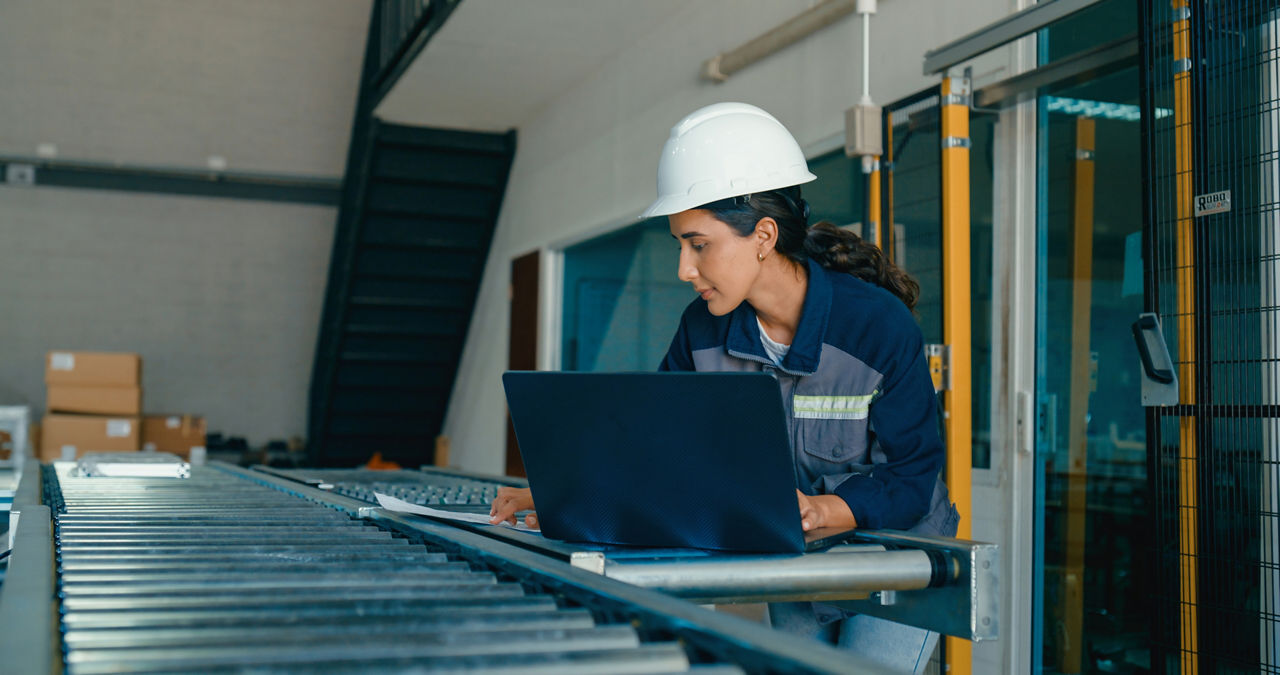 Young female engineer wearing hardhat uses laptop to analyze data and manage operations in factory. Robotic Industrial Factory Concept.