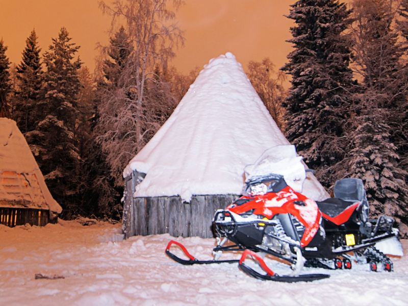 Vor zugeschneiten Waldhütten steht ein Schneemobil.