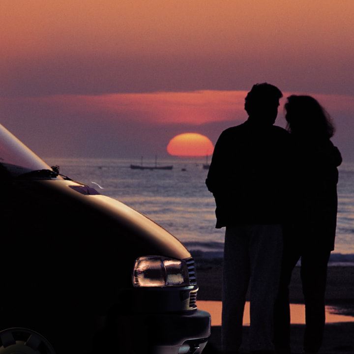 Ein Pärchen steht mit dem Volkswagen Nutzfahrzeuge California am Strand und betrachtet den Sonnenuntergang.
