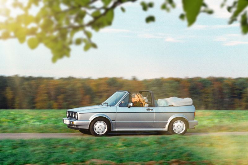 Two women drive a vintage Golf Cabrio along a country road.